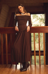 A woman is wearing an off-the-shoulder, long-sleeved, dark brown dress with a fitted bodice and a pleated, floor-length skirt. She accessorizes with a large, chunky silver necklace and a ring on her left hand. She is also wearing black heeled boots.
